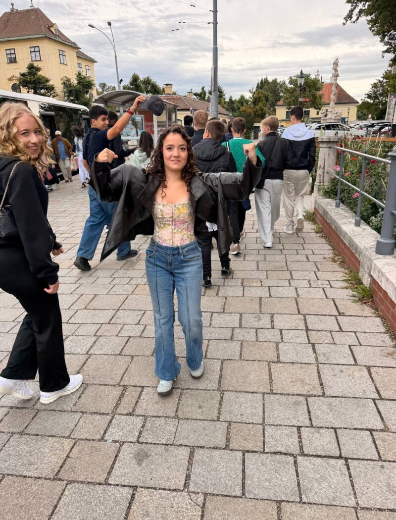 A student raises both arms playfully while classmates walk along a paved path near a bus stop.