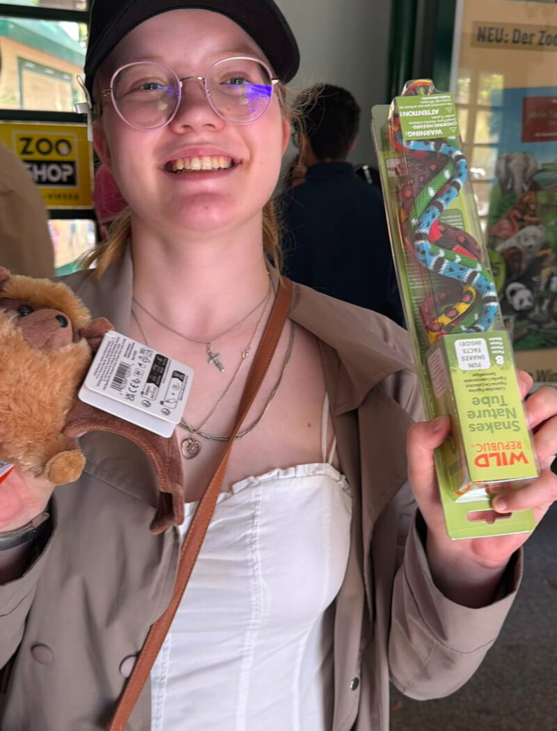 A smiling student wearing a black cap holds a plush animal and a tube of toy snakes in the zoo shop.
