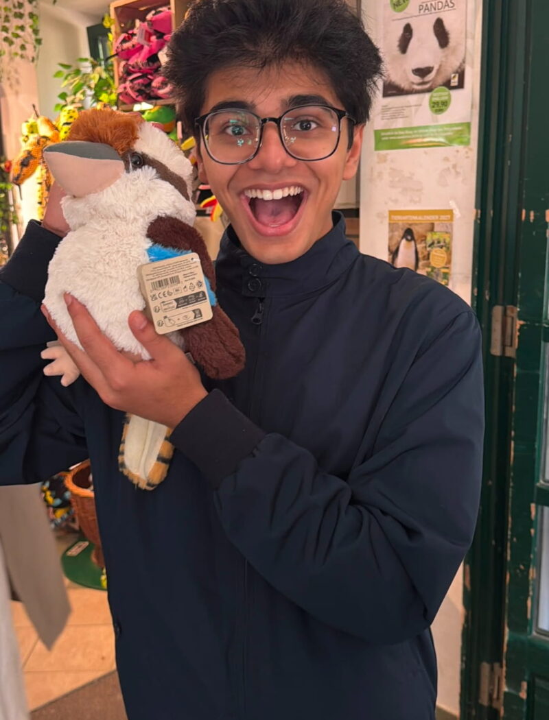 A student smiles while balancing a tall stack of plush animals on their head in a bright classroom.