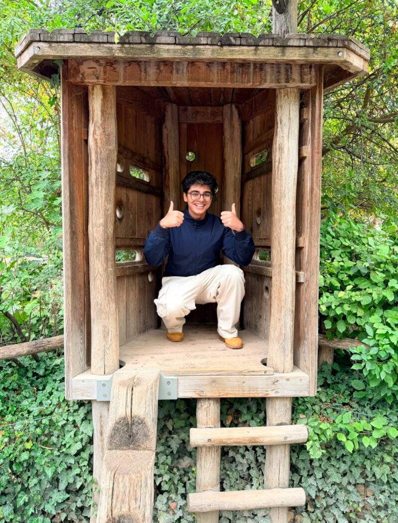 A student crouches inside a small wooden play tower and gives two thumbs up.