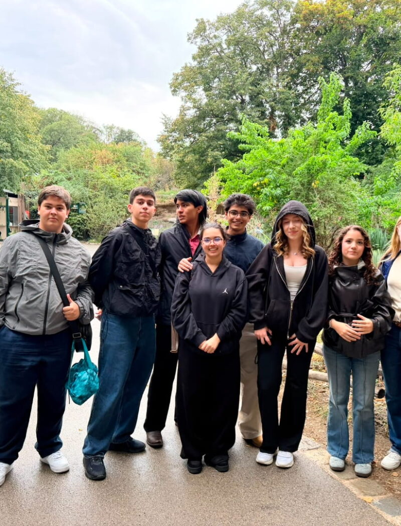 Nine students pose together on a zoo path surrounded by trees, plants, and wooden fencing.