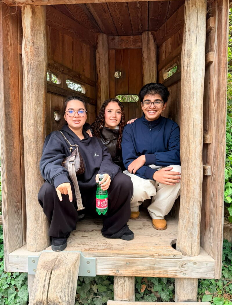 Three students pose together inside a small wooden play tower surrounded by greenery.