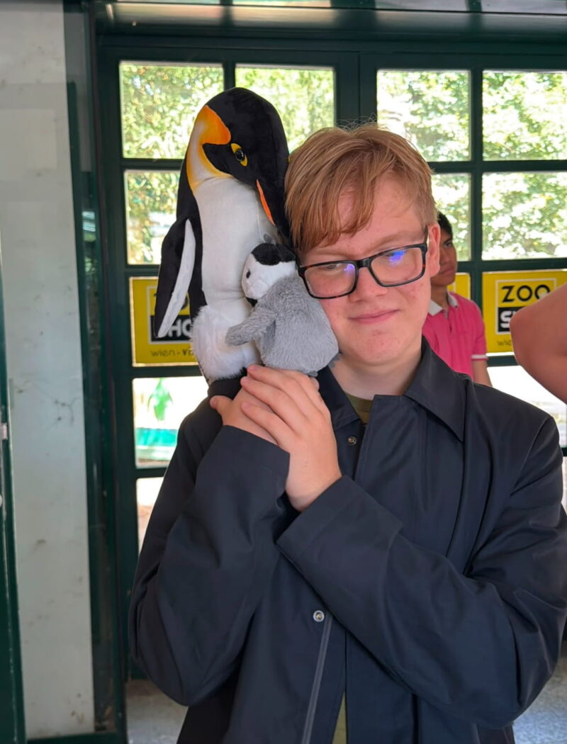 A student wearing glasses holds a large plush penguin and a smaller penguin chick against their shoulder.