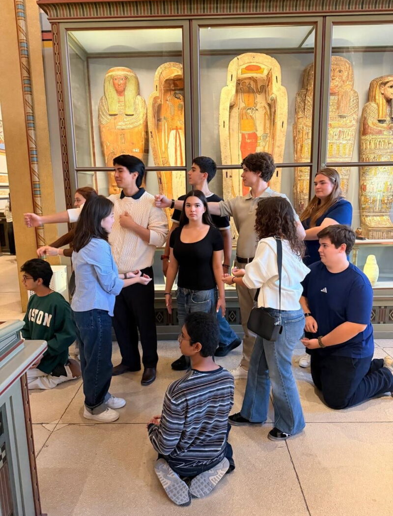 Students arrange themselves in playful poses in front of a museum display of decorated Egyptian coffins.