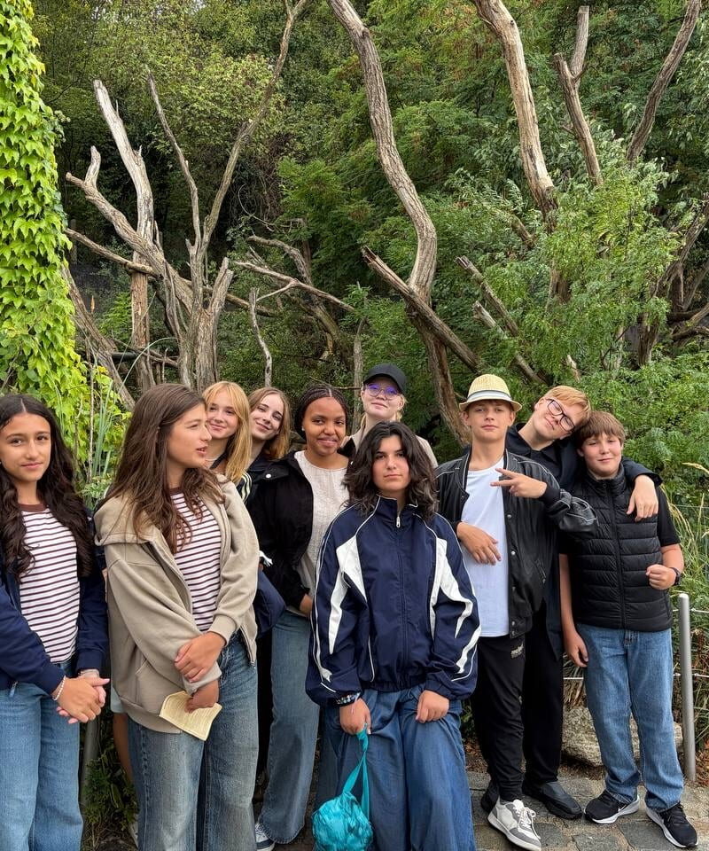 Students stand together in front of dense vegetation and tall bare branches at Schönbrunn Zoo.