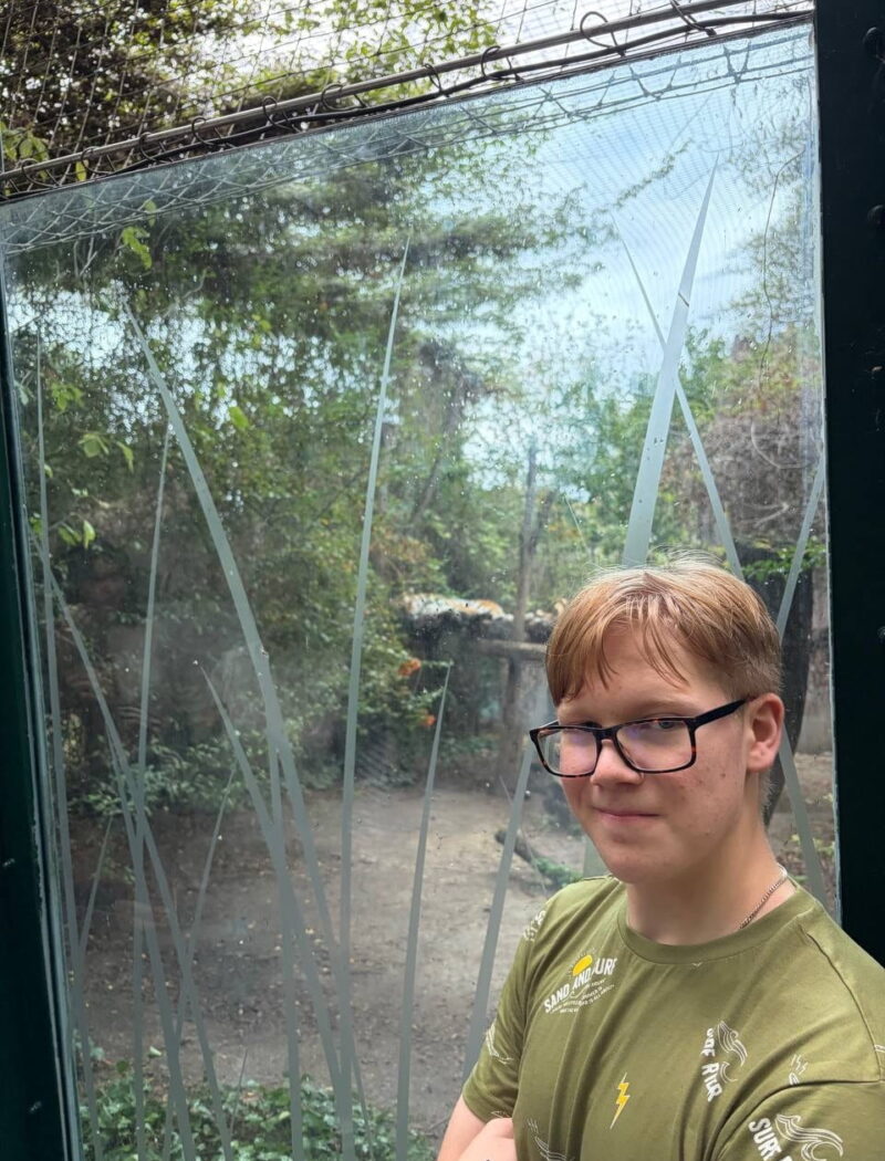 A student in glasses and a green T-shirt stands beside a glass viewing panel overlooking a leafy animal enclosure.
