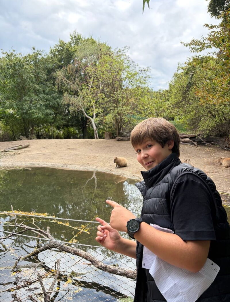 A student in a black padded vest points towards a capybara beside a pond in its enclosure.