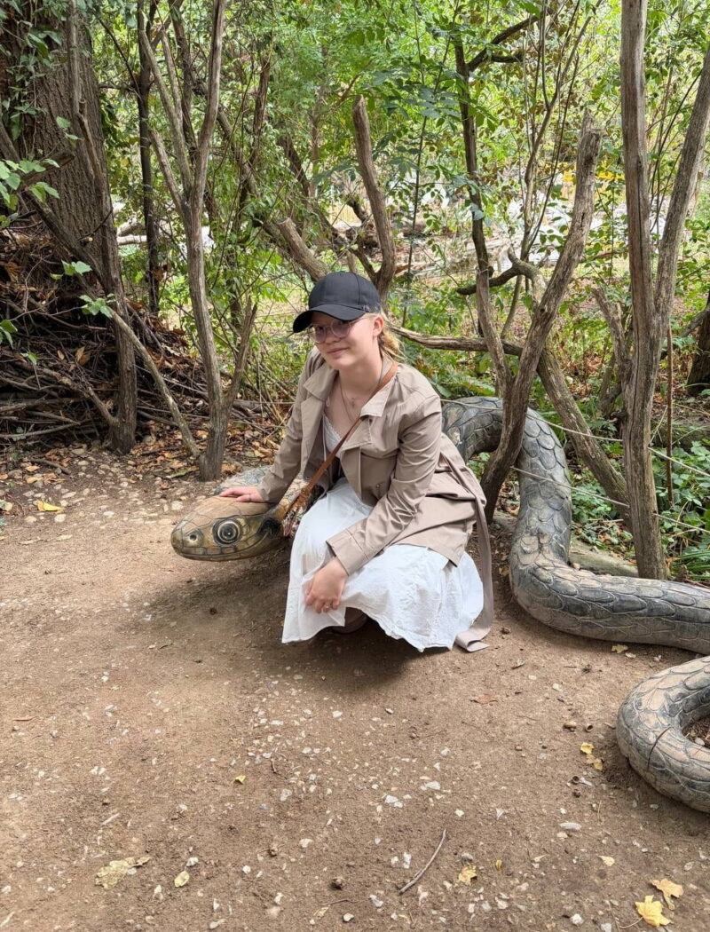 A student wearing a black cap and beige coat crouches beside a large snake sculpture among trees.