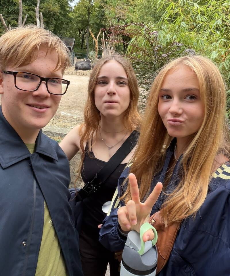 Three students take a selfie beside a leafy enclosure, with one student making a peace sign.