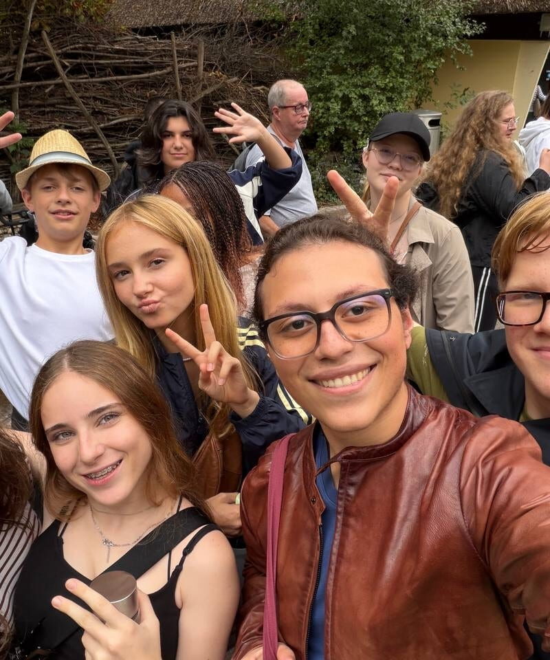 Students and a person in a brown leather jacket pose for a group selfie, with several students raising peace signs.