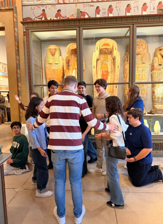 Our school group gathers in front of decorated Egyptian coffins, with several students kneeling for a group photo.