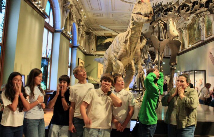 Schloss Krumbach International School students pose with playful expressions of surprise beside a large dinosaur model in a museum gallery.