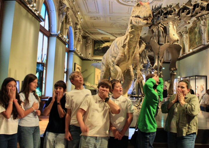 Schloss Krumbach International School students pose with playful expressions of surprise beside a large dinosaur model in a museum gallery.