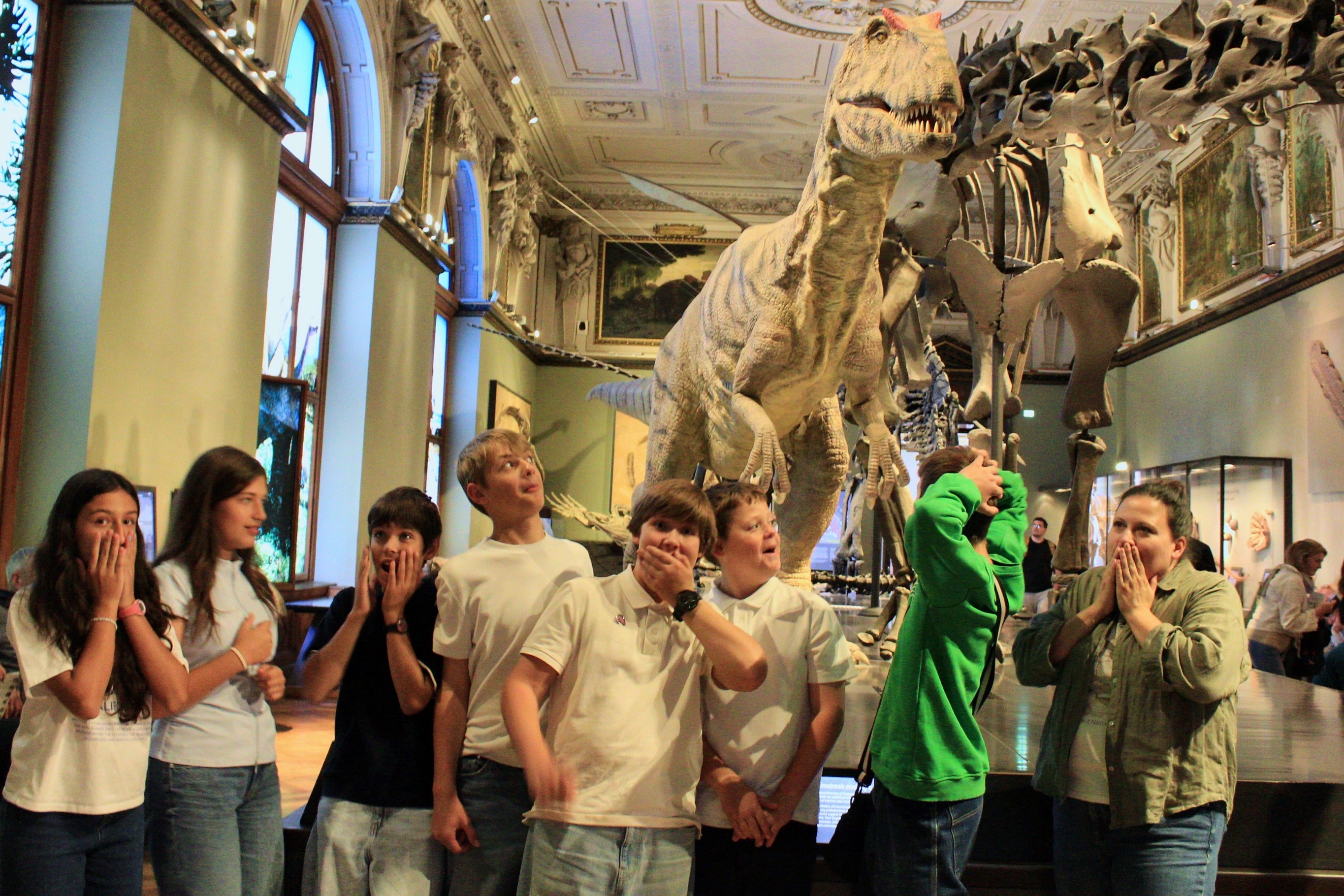Schloss Krumbach International School students pose with playful expressions of surprise beside a large dinosaur model in a museum gallery.