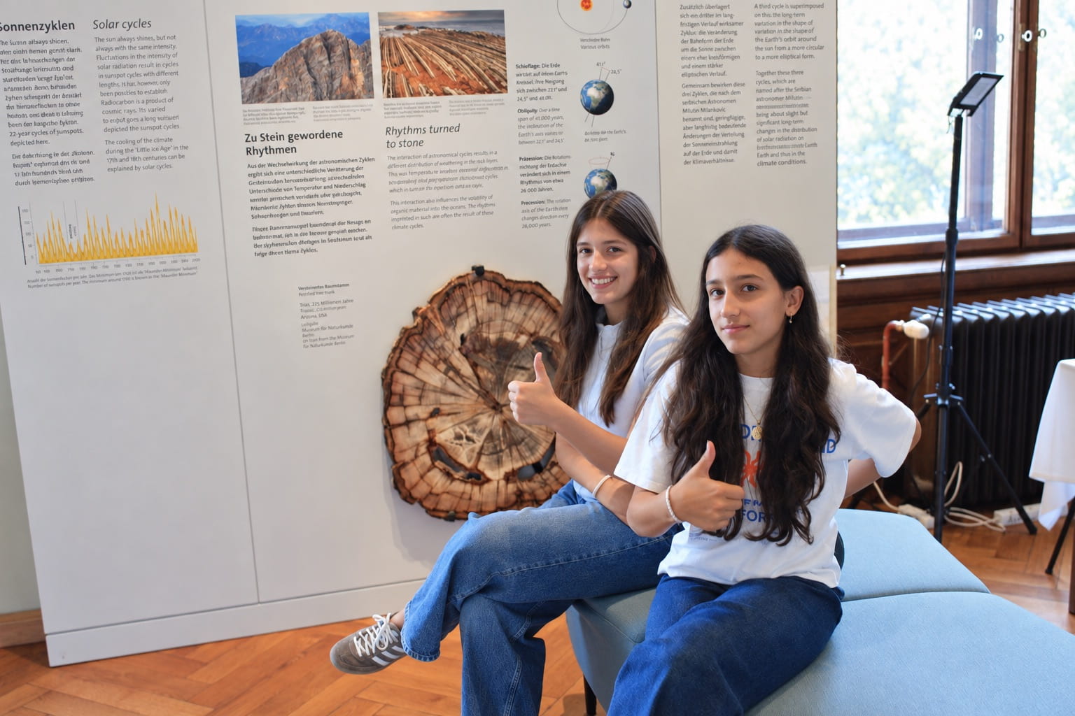 Two Schloss Krumbach International School students give thumbs up beside a petrified tree trunk display during a museum visit.