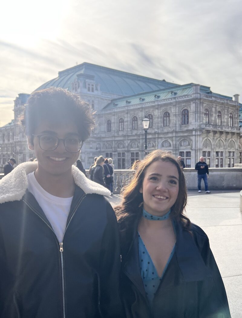 Two Schloss Krumbach International School IB Visual Arts students smiling outdoors with Vienna's historic State Opera House in the background.