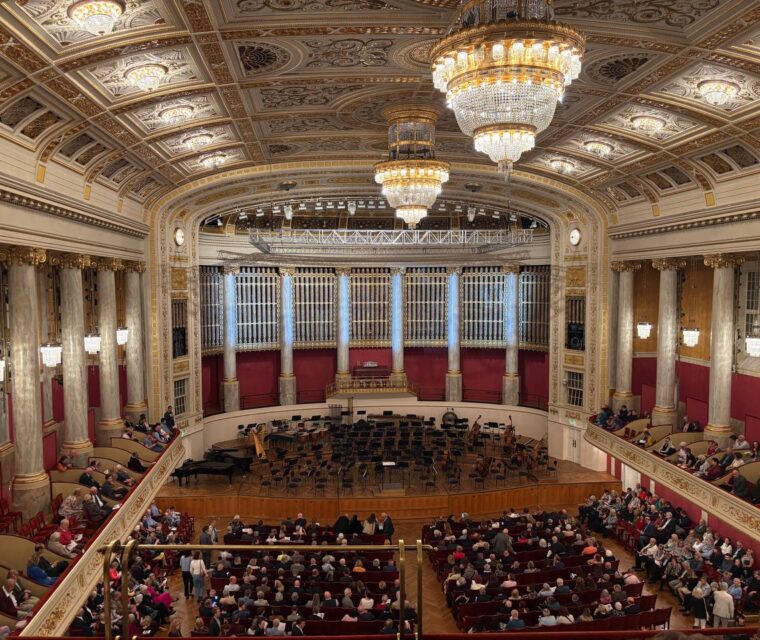 Interior of the Wiener Konzerthaus concert hall featuring ornate ceilings, crystal chandeliers, red auditorium seating, and an orchestra stage.